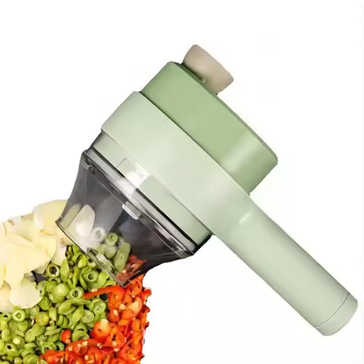 A light green manual food chopper with a clear plastic bowl and handle, shown cutting green beans and red chili peppers on a white background.